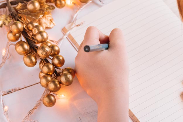 Blog vital: Cómo hacer propósitos alcanzables para el año nuevo, mano de persona escribiendo con un esfero sobre papel y al lado un ramillete de esferas de navidad color dorado