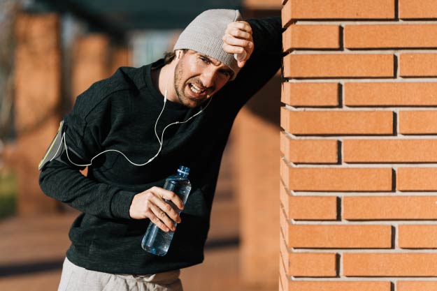 Hombre atlético agotado apoyado en una pared mientras toma agua después de hacer ejercicio al aire libre.